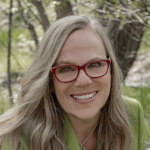 Photo of Sally Spencer Thomas smiling; she has blonde hair and is wearing a green jacket and red glasses.