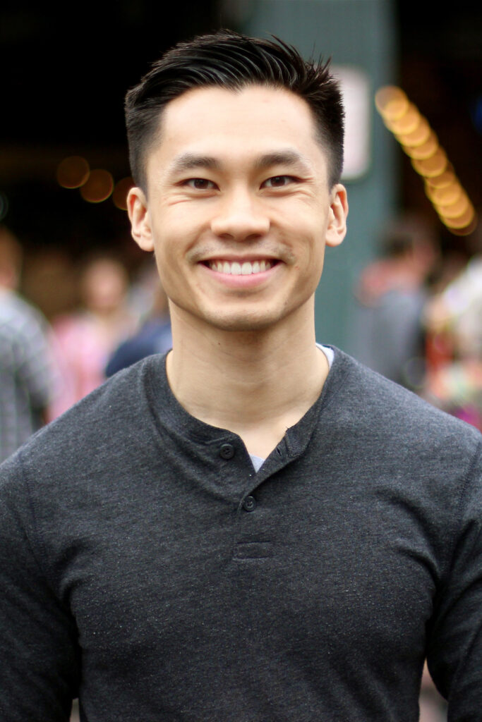 Hoan Do smiling with a blurred crowed of people walking behind him. He has short dark colored hair, and is wearing a dark grey henley style long sleeve shirt.